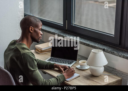 Vue de côté de l'accent african american man making notes dans l'ordinateur portable tout en restant assis à la maison travail de bureau Banque D'Images