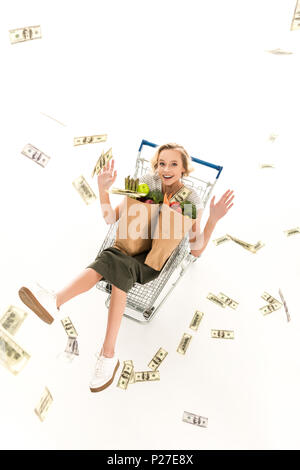 Excitée jeune femme avec des sacs d'épicerie en séance shopping trolley et jeter billets dollar isolated on white Banque D'Images