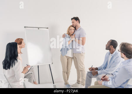 Middle aged couple holding hands while standing près de tableau blanc et d'autres gens assis sur des chaises au cours de la thérapie de groupe Banque D'Images