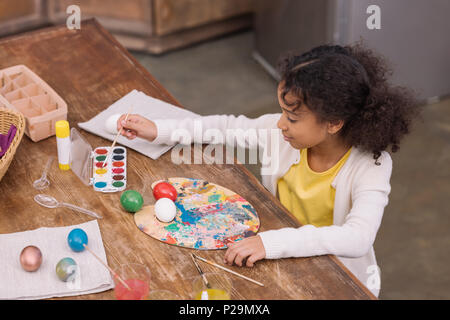 High angle view of african american kid mettre brosse dans les peintures à l'aquarelle pour peindre les oeufs de pâques Banque D'Images