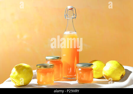 Gelée de coing faite maison et le jus dans une bouteille et en pots de verre avec coings sur une nappe de lin en plein soleil devant un fond orange et jaune Banque D'Images