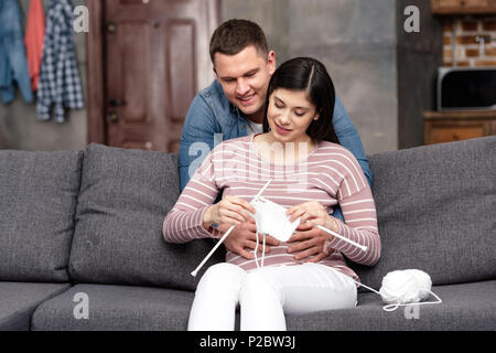 Smiling young man hugging pregnant woman sitting on sofa et tricoter à la maison Banque D'Images