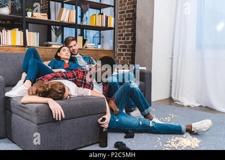 Les jeunes amis multiethnique de dormir dans la chambre en désordre avec pop-corn et les bouteilles de bière Banque D'Images