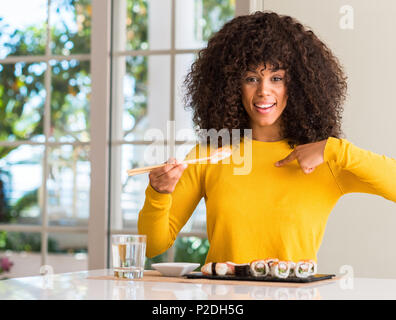 African American Woman eating sushi à l'aide de baguettes à la maison avec surprise face à face à lui-même Banque D'Images