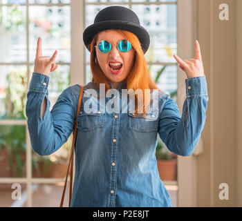 Rousse élégante femme portant chapeau melon et des lunettes en criant avec crazy expression faisant rock symbole avec les mains. Music star. Concept lourd. Banque D'Images