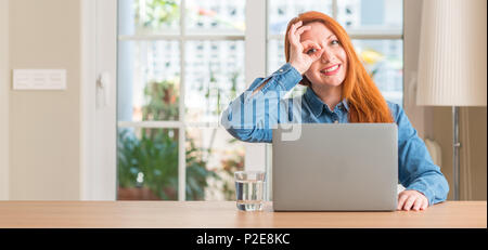 Femme rousse à l'aide d'un ordinateur ordinateur portable à la maison avec happy smiling face faire ok signe avec la main sur de l'oeil Banque D'Images