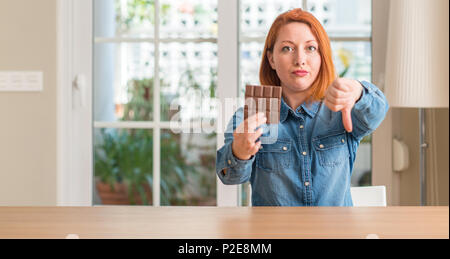 Redhead woman holding barre de chocolat à la maison, visage en colère avec signe négatif montrant non avec vers le bas, le rejet de la notion Banque D'Images