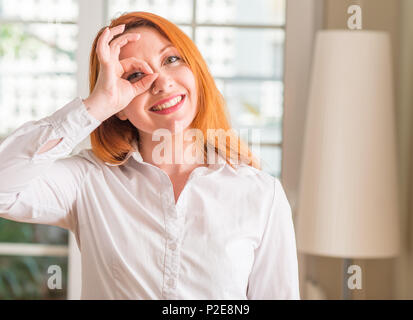 Redhead woman wearing white shirt à la maison avec happy face smiling faire ok signe avec la main sur de l'oeil Banque D'Images