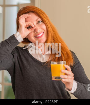 Redhead woman holding orange verre de jus à la maison, avec happy smiling face ok faire signe avec la main sur de l'oeil Banque D'Images