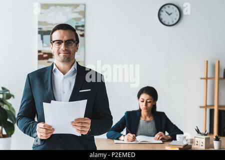 Businessman holding papers tandis que femme travaillant par table avec bloc-notes de bureau léger Banque D'Images