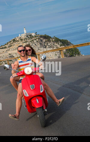 Jeune couple riding a scooter Vespa rouge sur route le long de la péninsule de Cap de Formentor avec Faro de Formentor lighthouse derrière, Palma Banque D'Images