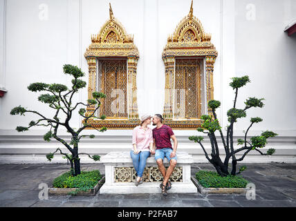 Jeune couple en habits rouges assis sur le banc près d'arbres décoratifs et golden windows de Wat Pho à Bangkok, Thaïlande Banque D'Images