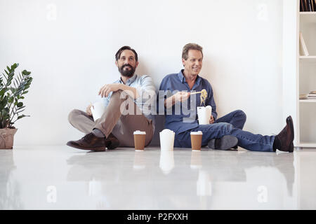 Deux hommes d'âge moyen de manger des aliments asiatiques et de boire du café tout en étant assis sur le plancher in office Banque D'Images