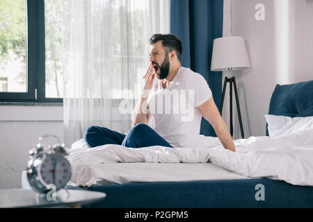 Jeune homme barbu en pyjama le bâillement tandis qu'assis sur le lit, le matin Banque D'Images