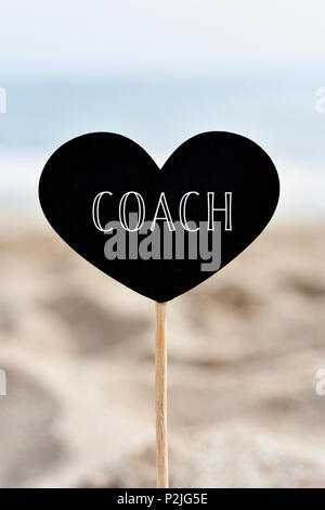 Libre d'une pancarte en forme de coeur noir avec texte coach écrit en C, attaché à un bâton, sur le sable d'une plage, avec la mer en arrière-plan Banque D'Images