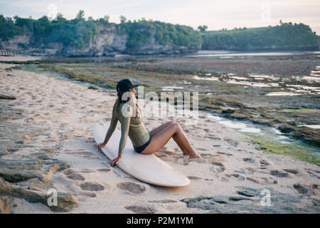 Jeune femme séduisante en wetsuit assis sur une planche de surf sur le littoral et à la recherche au coucher du soleil Banque D'Images