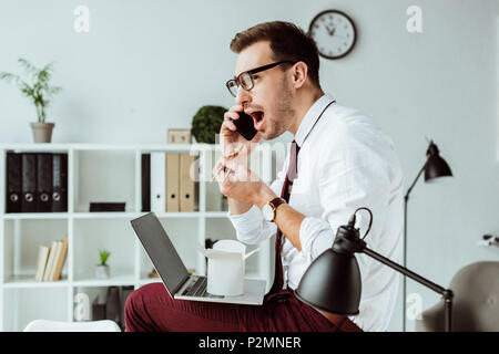 Man avec le smartphone et l'ordinateur portable tout en mangeant des nouilles à emporter pour le déjeuner in office Banque D'Images