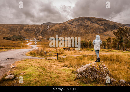 Un female hiker debout sur un rocher au milieu de l'herbe jaune, regard vers les montagnes au loin, contre un ciel sombre et moody à Glencoe, Sco Banque D'Images