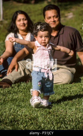 Les premiers pas de bébé à l'extérieur de l'écart des problèmes de mise au point les parents assis sur l'herbe. De descendance indienne M. © Myrleen frontale ....Pearson Ferguson Cate Banque D'Images