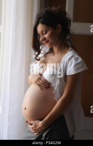 Femme enceinte de sept mois est par la fenêtre avec ses mains autour de son ventre et une expression sereine sur son visage, perdu dans la pensée de l'avenir. Banque D'Images