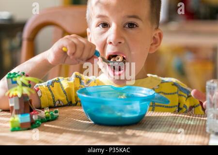 Le garçon 4 ans mange du porridge. Table enfant. Le concept de l'indépendance de l'enfant. Le garçon est déjeunant avec un appétit sur l'arrière cuisine Banque D'Images