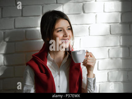 Pretty smiling student boire du café le matin. Avoir les cheveux noirs courts et lumière jour composent. Portant une chemise blanche et un cardigan rouge. Se sentir bien, heureux. Mur Blanc arrière-plan. Banque D'Images