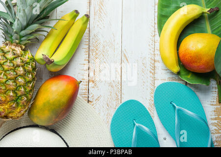 Mise à plat des fruits tropicaux Composition Arrangement Mangue Ananas Banane Feuille de palmier vert Chapeau femme chaussons bleu sur fond de bois Revêtus de blanc. Vac Banque D'Images