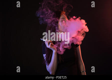 Jeune femme fumant cigarette électronique sur fond sombre Banque D'Images