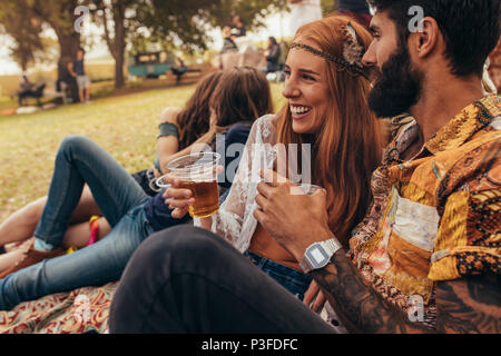 Smiling couple avec beers assis dans un parc et profiter du festival d'été. Heureux l'homme et de la femme avec des amis en arrière-plan. Banque D'Images