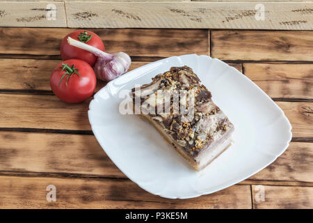 Un morceau de viande de porc se trouve sur une plaque carrée blanche. Sur la surface en bois de la table, les tomates et l'ail Banque D'Images