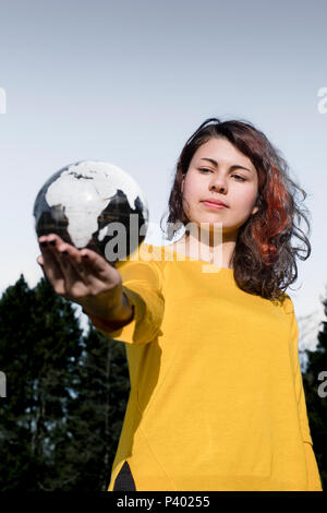 Le monde entier dans sa main : young woman in stylish pull jaune tenant dans sa main la terre en face de ciel et les arbres. Sauver la Terre concept. Banque D'Images