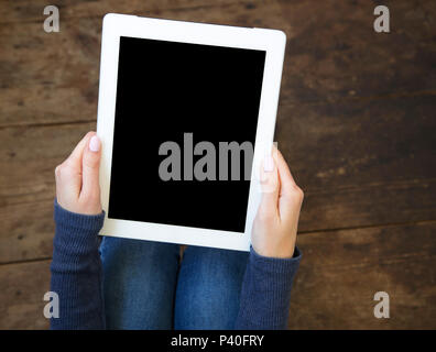 La jeune fille est assise sur un sol en parquet et est titulaire d'une tablette avec un écran vide dans ses mains. Vue d'en haut. Banque D'Images