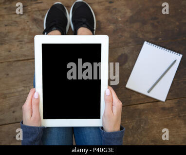 La jeune fille est assise sur un sol en parquet et est titulaire d'une tablette avec un écran vide dans ses mains. Ordinateur portable sur le sol. Vue d'en haut. Banque D'Images