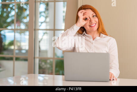 Femme rousse à l'aide d'un ordinateur ordinateur portable à la maison avec happy smiling face faire ok signe avec la main sur de l'oeil Banque D'Images