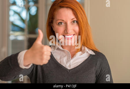 Femme rousse à la maison heureux avec un grand sourire faisant signe ok, pouce vers le haut avec les doigts, signe d'excellentes Banque D'Images