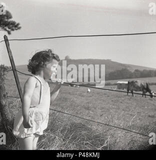 Historique années 1950, photo d'un bébé fille à l'extérieur dans la campagne holding un fil d'une vieille clôture en regardant les vaches dans le champ, England, UK. Banque D'Images