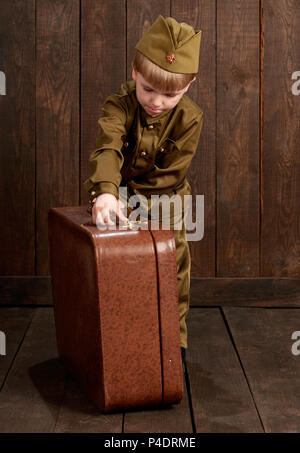 Les enfants garçon sont habillés en soldat en uniforme militaire rétro avec vieille valise, fond en bois foncé, style retro Banque D'Images