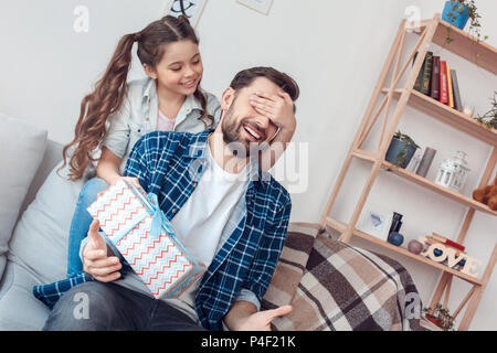 Homme barbu et petite fille à l'accueil en famille assis sur canapé fille fermer les yeux du père présent donnant fort surprise faisant sourire enjoué Banque D'Images