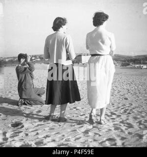 Années 50, deux dames debout ensemble sur une plage de sable fin, tandis qu'un gentilhomme à genoux prend une photo avec son appareil photo (ci-dessous), Angleterre, Royaume-Uni. Banque D'Images