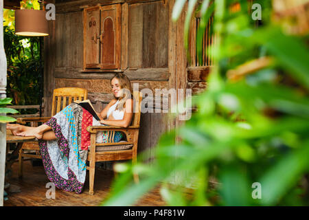 Jolie jeune fille s'asseoir sur la véranda en plein air de bungalow en bois avec vue sur le jardin tropical, lisez romance au livre papier. Young woman relaxing in Luxury villa Banque D'Images