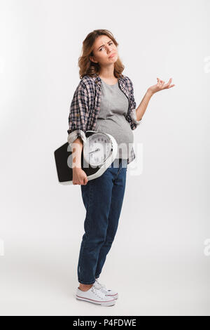 Contrarié young pregnant woman holding échelles et looking at camera isolated on white Banque D'Images