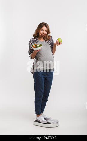 Smiling pregnant woman avec chocolat, salade de légumes et d'Apple sur les balances permanent isolated on white Banque D'Images