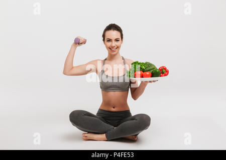 Happy young woman sitting on floor avec bac de divers légumes sains et montrant ses muscles isolated on white Banque D'Images