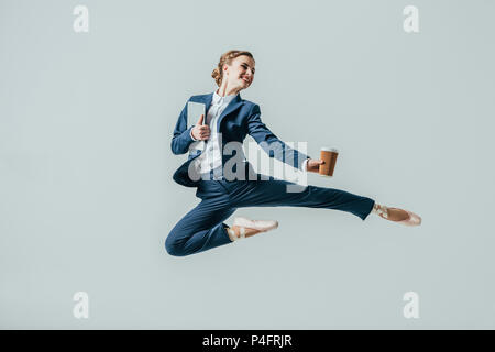 Businesswoman in suit et chaussures de ballet sautant avec café et tablette numérique, isolé sur gris Banque D'Images