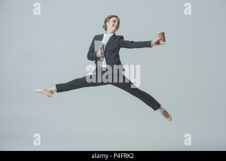 Businesswoman in suit et chaussures de ballet sautant avec café et comprimé, isolé sur gray Banque D'Images