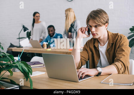 Businessman using laptop and talking on smartphone et collègues qui travaillent derrière in modern office Banque D'Images