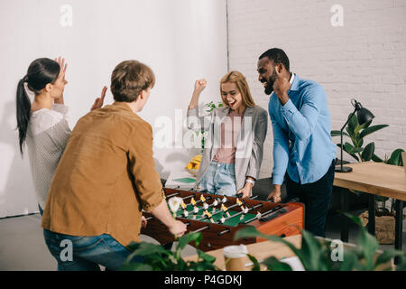 Heureux collègues d'entreprises multiculturelles gesticulant et playing table football in modern office Banque D'Images