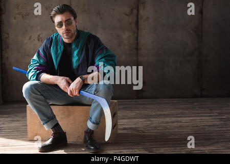 Beau jeune homme en vintage windcheater avec bâton de hockey assis sur boîte en bois Banque D'Images