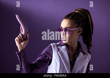 Élégant belle african american girl en violet lunettes holding banana comme arme, isolé sur purple Banque D'Images