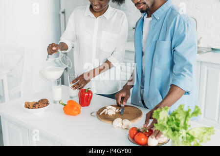 Vue partielle de l'african american woman pouring milk into glass tout en mari la cuisson petit déjeuner en cuisine à la maison Banque D'Images
